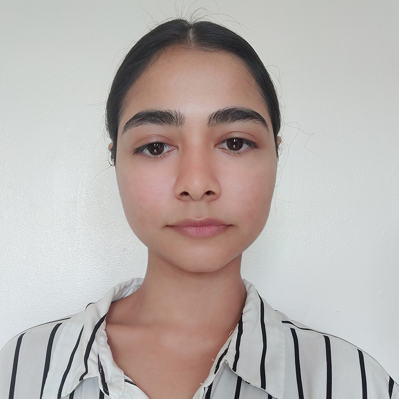 A woman with dark hair tied back, wearing a white shirt with black vertical stripes, stands against a plain white background, looking directly at the camera with a neutral expression.