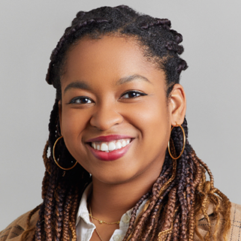 A woman with braided hair smiles at the camera, wearing gold hoop earrings, layered necklaces, a white collared shirt, and a tan sweater. She embodies the spirit of PEN Across America against a plain, light gray background.