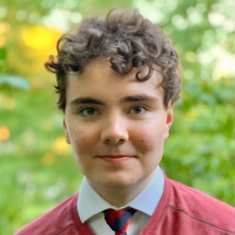 A young person with short curly hair, wearing a red sweater over a collared shirt and striped tie, stands outdoors with green foliage and sunlight blurred in the background.