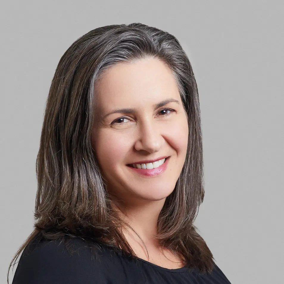 A woman with shoulder-length brown and gray hair smiles at the camera. She is wearing a black top and is posed in front of a plain gray background.