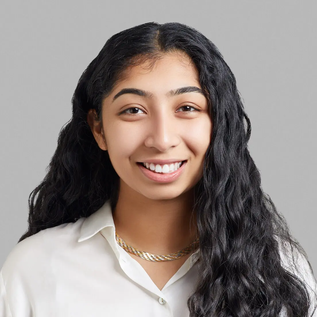 A young woman with long, wavy black hair smiles at the camera. She is wearing a white collared shirt and a gold chain necklace, standing against a plain gray background at the NYC Free Expression Advocacy Institute by PEN.