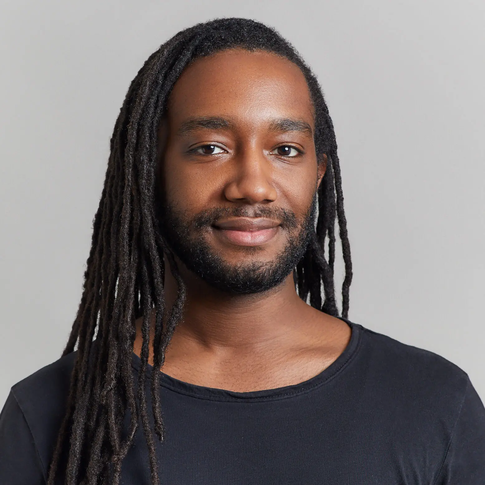 A person with long dark dreadlocks and a beard, wearing a black shirt, smiles softly while facing the camera against a plain light gray background, evoking themes of justice and resilience beyond prison walls.