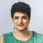 A person with short curly hair and a nose ring smiles at the camera. Wearing a green top, dangly earrings, and a necklace against a plain light background, they promote digital safety and awareness about online abuse.