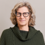 A middle-aged woman with wavy gray hair, wearing black-rimmed glasses and a dark green, ribbed sweater with a cowl neck, smiles softly at the camera against a plain light background.