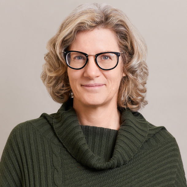 A middle-aged woman with wavy gray hair, wearing black-rimmed glasses and a dark green, ribbed sweater with a cowl neck, smiles softly at the camera against a plain light background.
