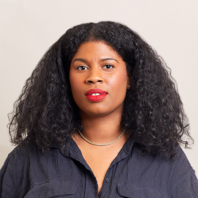 A woman with curly dark hair, wearing a dark textured shirt, a silver necklace, and red lipstick, stands against a plain light background, looking directly at the camera with a neutral expression.