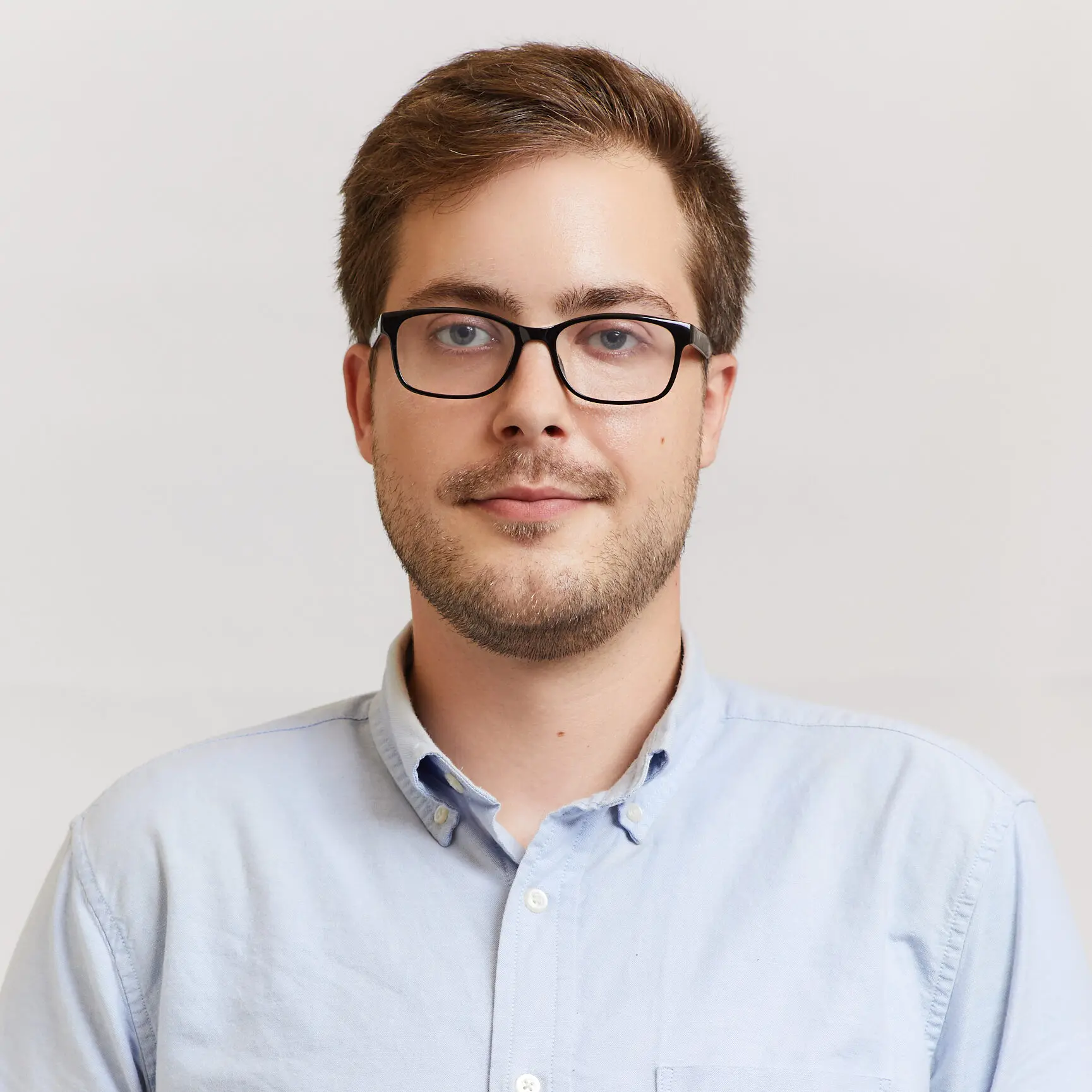 A young man with short brown hair, glasses, and light facial hair is wearing a light blue button-up shirt and standing against a plain, light background.