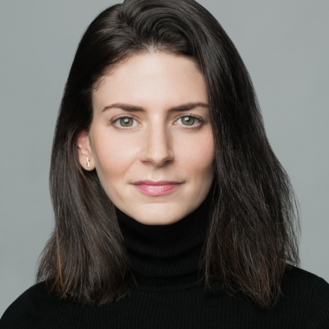 A woman with straight, shoulder-length dark hair and light eyes, wearing a black turtleneck sweater, looks directly at the camera against a plain gray background.
