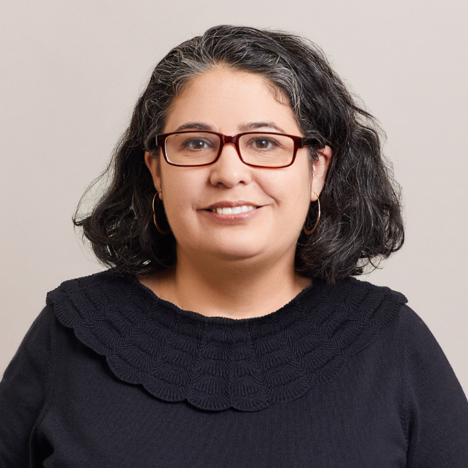 A woman with wavy, shoulder-length dark hair, wearing glasses, hoop earrings, and a black top with a scalloped collar, smiles at the camera against a plain light background.