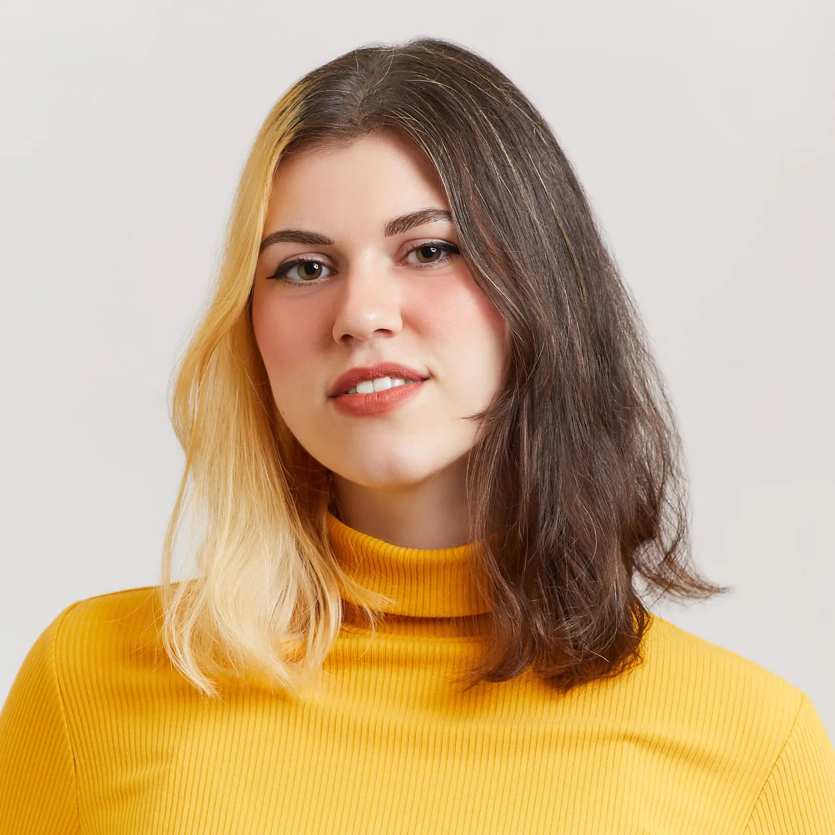 A young woman with shoulder-length hair, half dyed blonde and half brown, wearing a yellow turtleneck top, looks at the camera with a neutral expression against a light background.