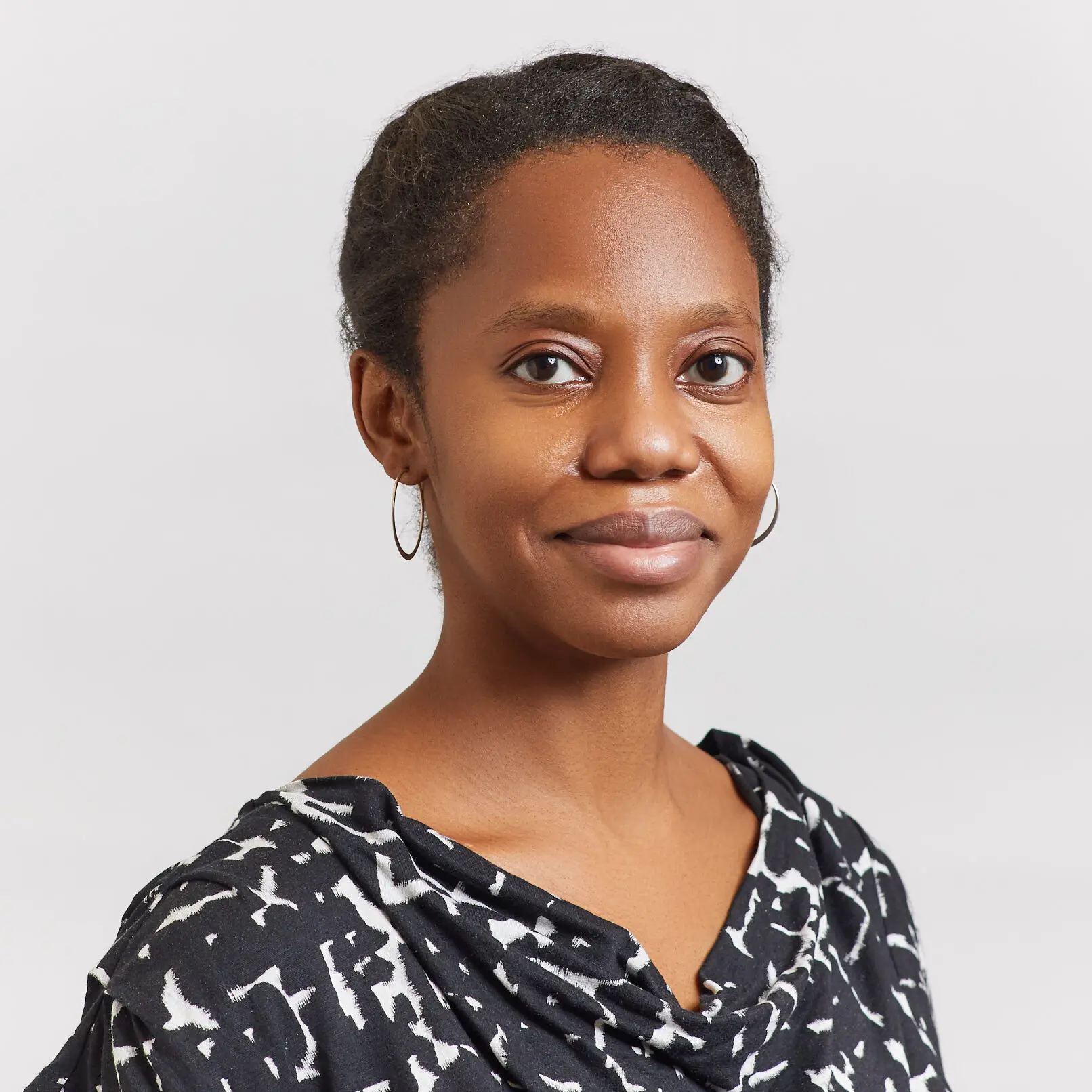 A woman with dark skin and short, natural hair is wearing hoop earrings and a black-and-white patterned top, smiling slightly, against a plain light background.