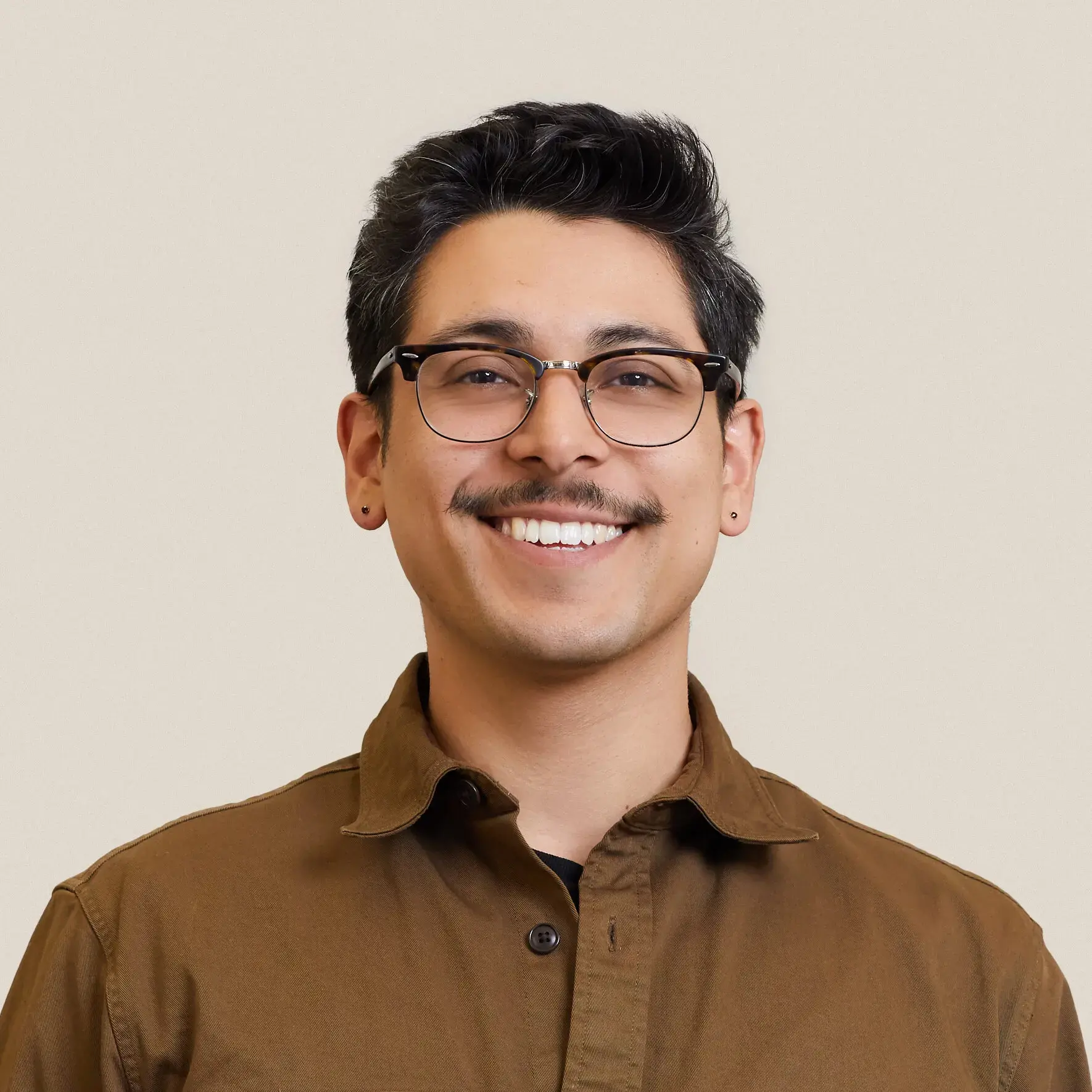 A smiling person with short dark hair, glasses, a mustache, and earrings, wearing a brown button-up shirt, standing against a plain light background.