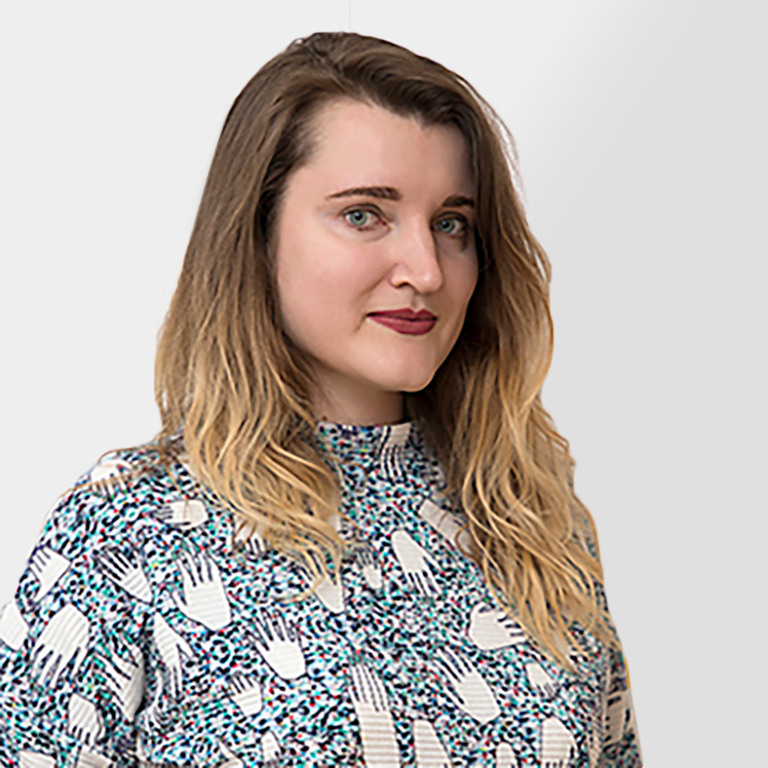 A woman with long, wavy blonde hair and fair skin, wearing a patterned top with blue, white, and black designs, stands in front of a plain light gray background, looking slightly to the side and smiling subtly.