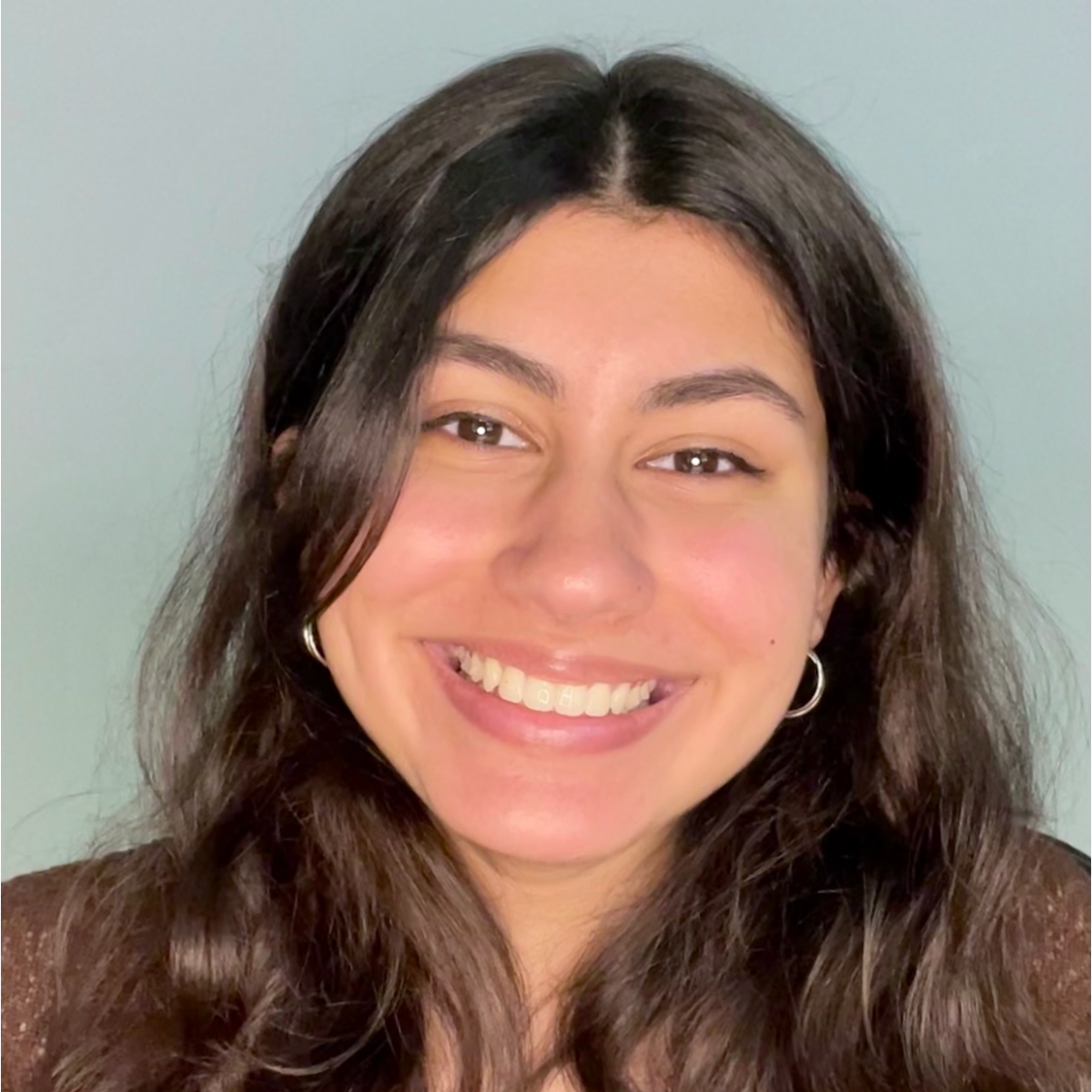 A young woman with long, dark wavy hair and hoop earrings smiles in front of a plain light blue background.