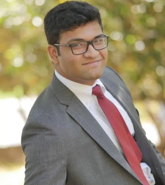 A young man wearing glasses, a gray suit, white shirt, and red tie smiles confidently while standing outdoors, with a blurred background of greenery and sunlight.