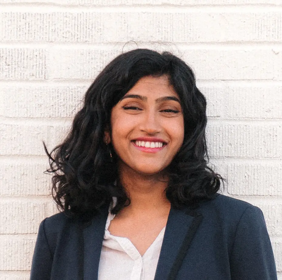 A woman with dark, wavy hair smiles while standing in front of a white brick wall. She is wearing a navy blazer over a white blouse.