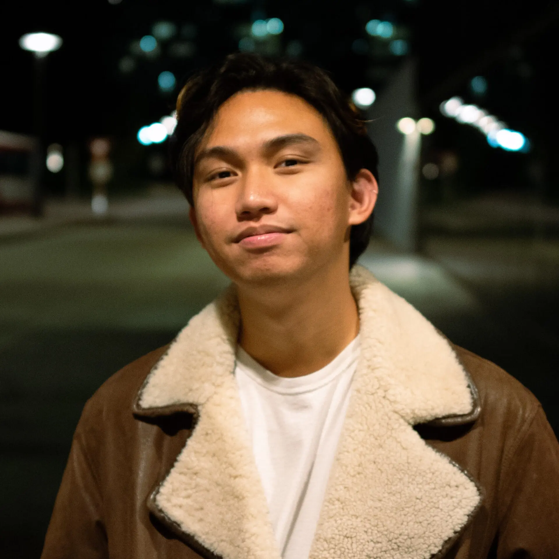 A young person with medium-length dark hair wears a brown shearling jacket over a white shirt, standing outdoors at night with blurred city lights in the background.
