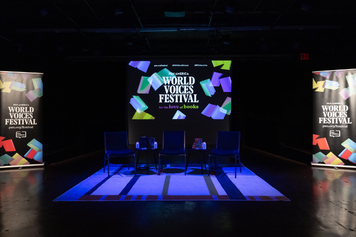 A stage setup with four empty chairs and a small table, illuminated by blue light, displays a screen reading World Voices Festival for the love of books with colorful book graphics and banners on each side.