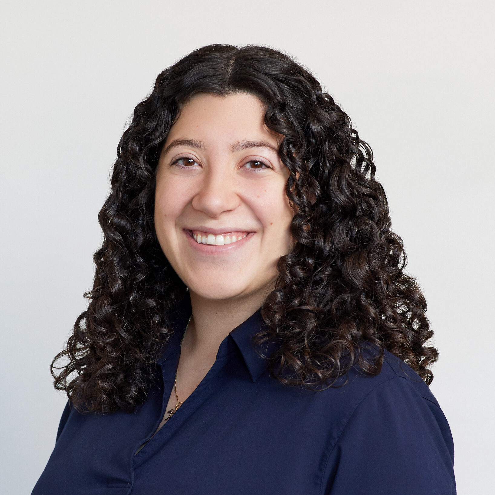 A woman with long, dark, curly hair smiles at the camera. She is wearing a dark navy blouse and is posed in front of a plain, light-colored background.