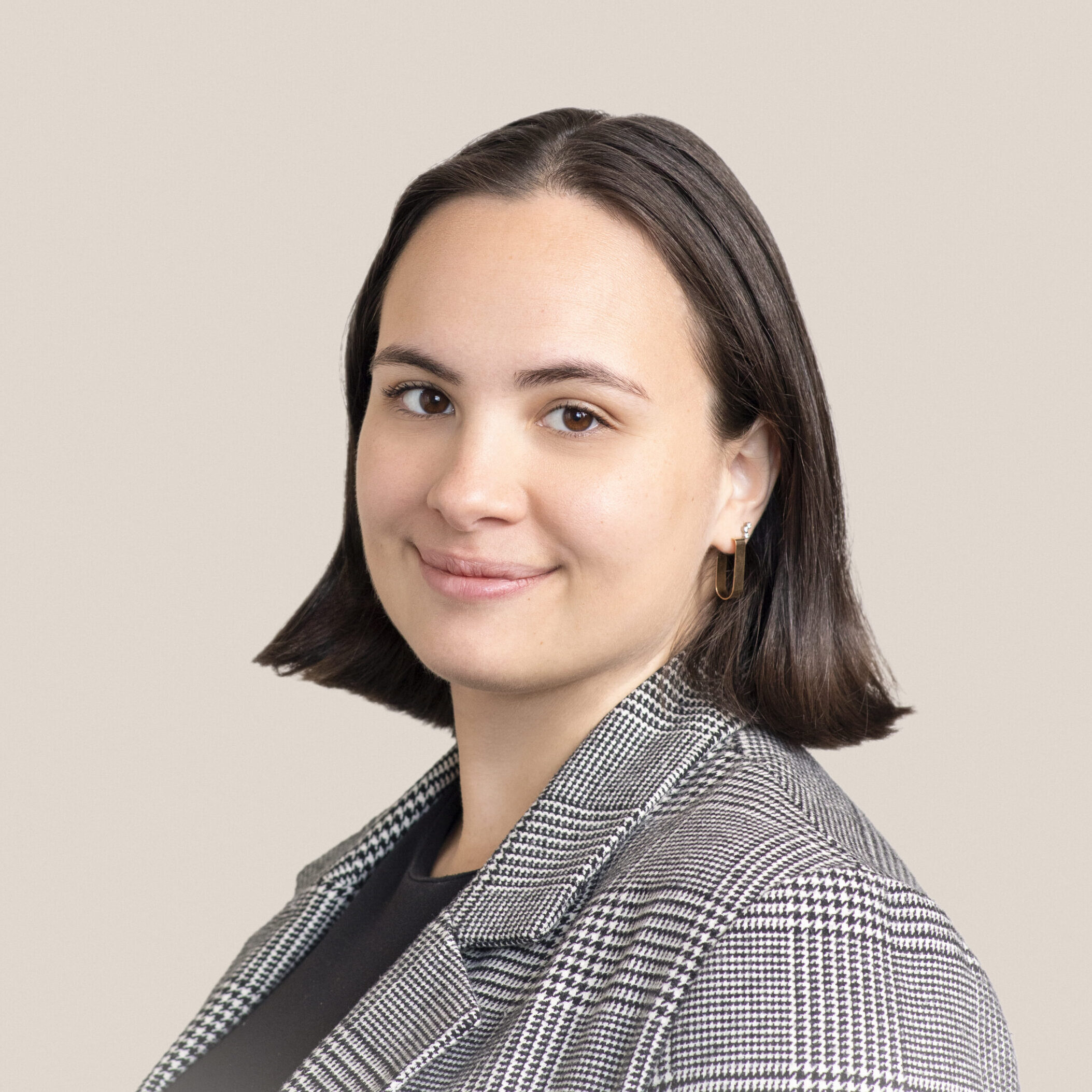A person with shoulder-length dark hair is smiling slightly. They are wearing a plaid blazer over a dark top. The background is a solid, light beige color.