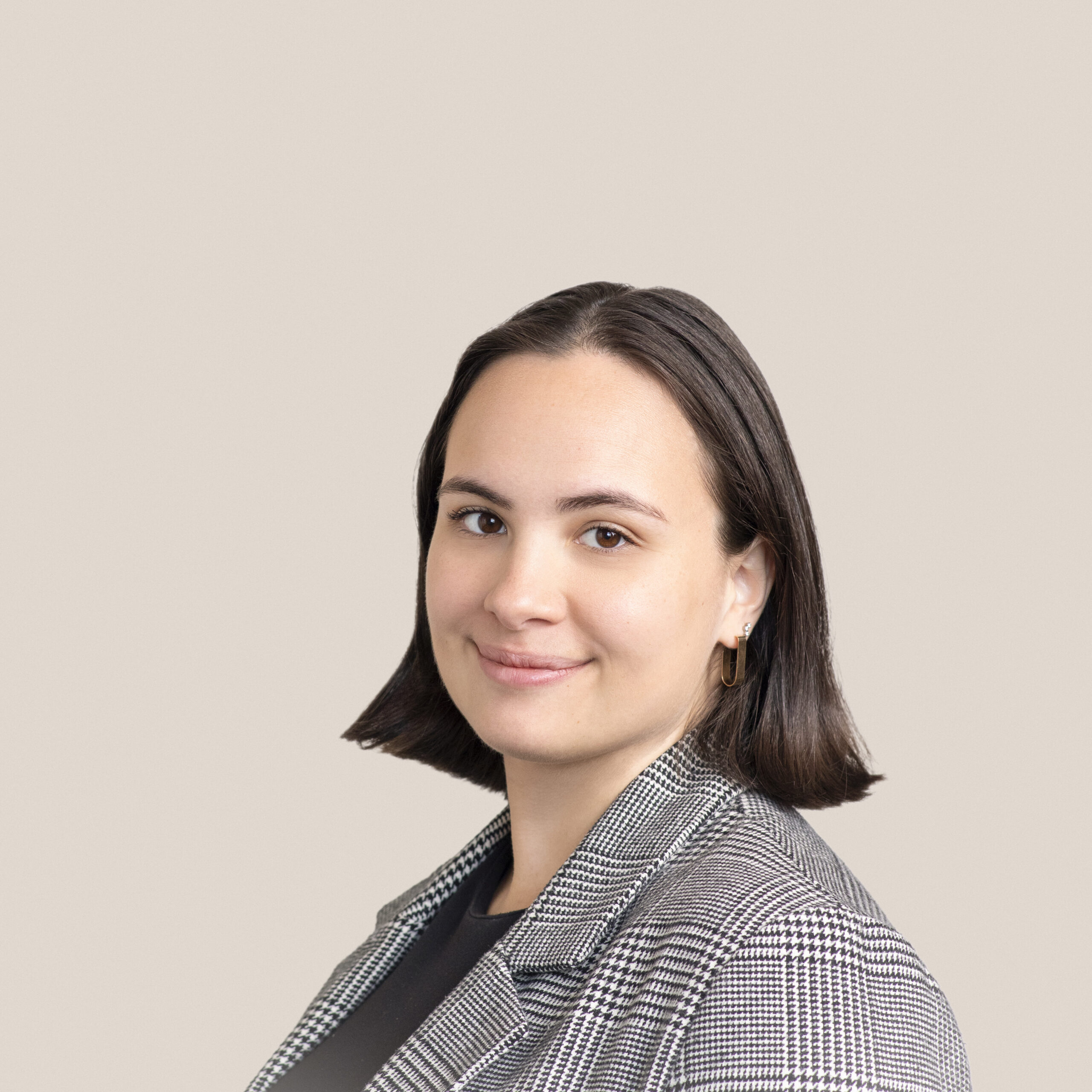 A person with shoulder-length dark hair is smiling slightly. They are wearing a plaid blazer over a dark top. The background is a solid, light beige color.