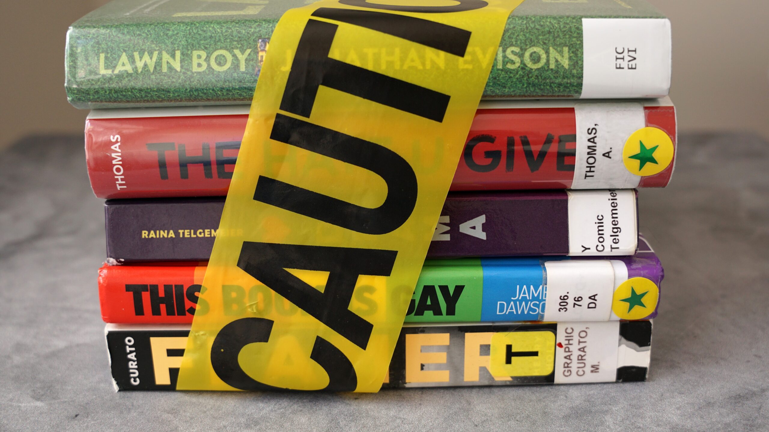 A stack of colorful books is partially covered by yellow caution tape, hinting at U.S. Free Expression Issues. The book spines show library labels and star stickers, with some titles visible but partially obscured by the tape.