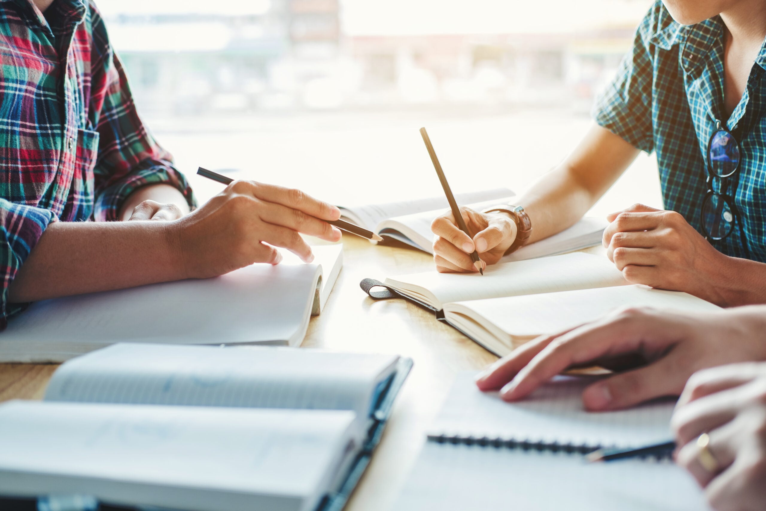 Three people sit at a table for a collaborative study session, writing in notebooks and reading books—only their hands and arms visible. The scene, reminiscent of Pen America workshops, is brightened by natural light from the background.
