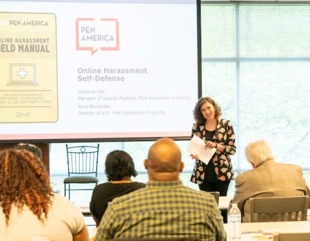 A woman stands at the front of a room giving a presentation titled Online Harassment Self-Defense to an audience, as part of PEN America workshops. A screen displays the PEN America logo and a manual cover.