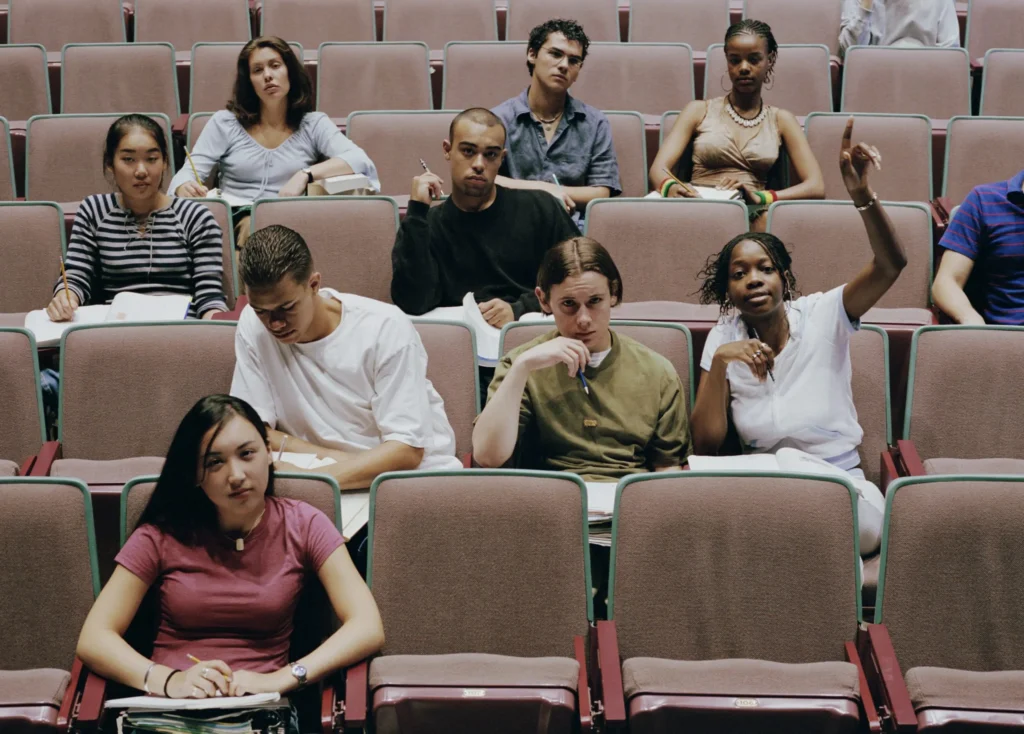 A diverse group of students sits in an auditorium, engaged and holding notebooks. One student in the front raises a hand, indicating participation in a discussion about educational censorship. The setting suggests a lecture or classroom environment.