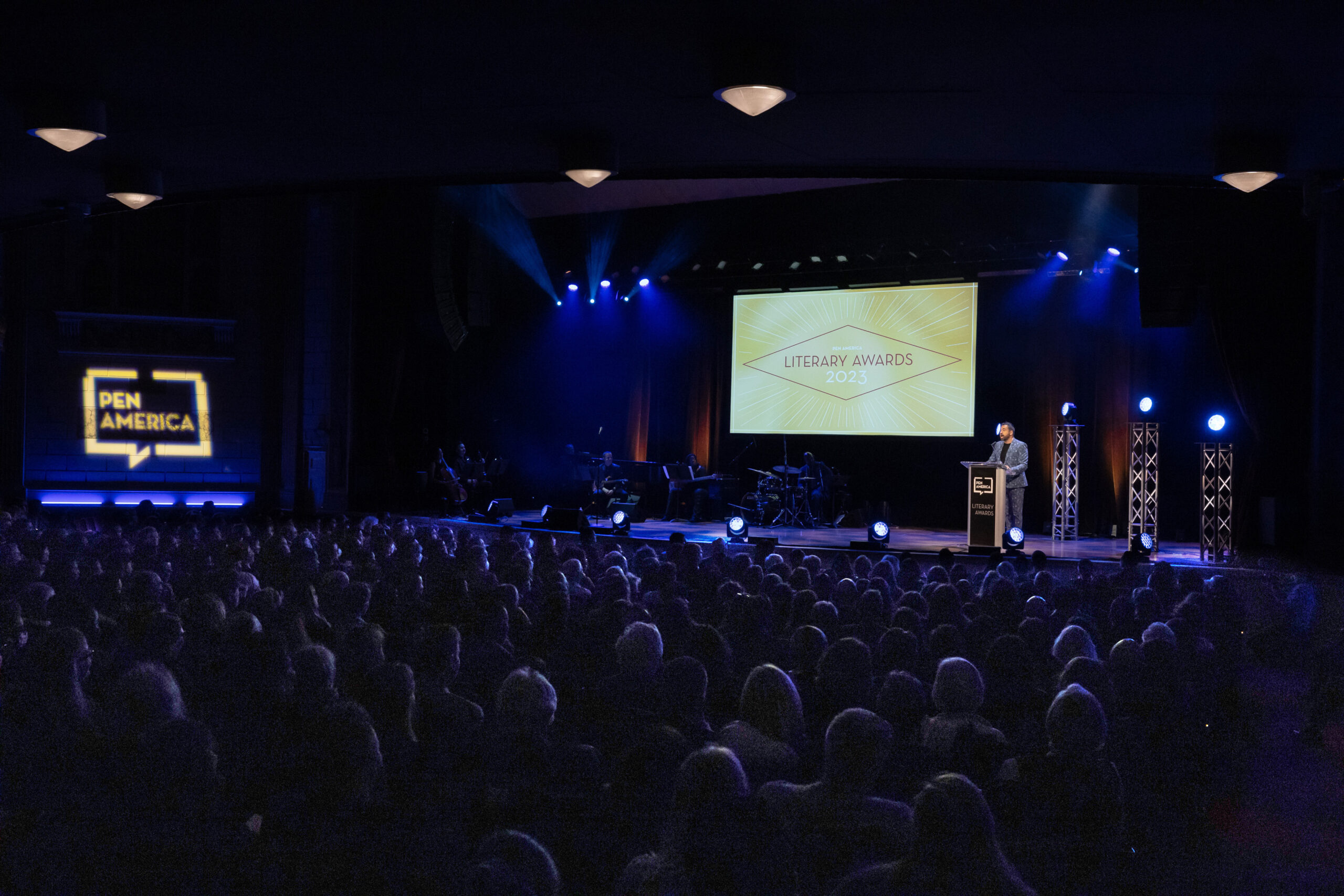 A person stands at a podium on stage during the 2021 PEN America Literary Awards. The audience is seated in a dimly lit theater. Blue and orange stage lights illuminate the stage, and a large screen displays the event name.