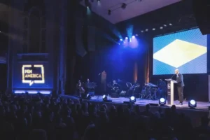 A person speaks at a lectern on a stage at a PEN America event. The room is dimly lit with blue and purple lights. A large screen displays a geometric design, and a band is performing on the side. An audience is seated in front.