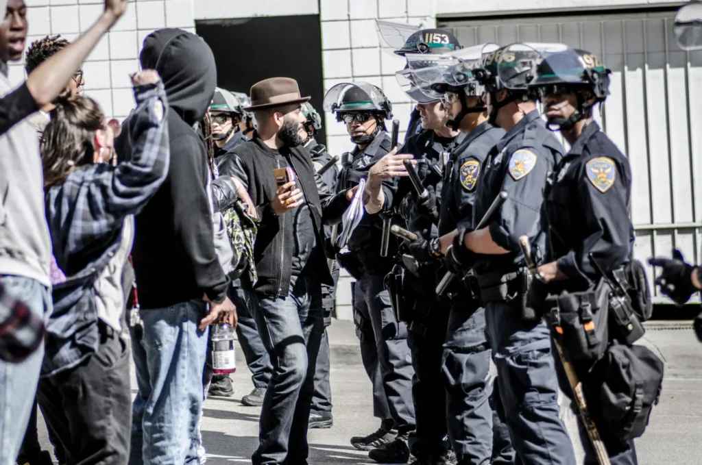 Protesters face a line of police officers in riot gear on a street, embodying the California Effect. One person raises their fist, while another captures the moment on a smartphone. Officers stand in line, some gesturing toward the crowd against a backdrop featuring a building with a garage door.