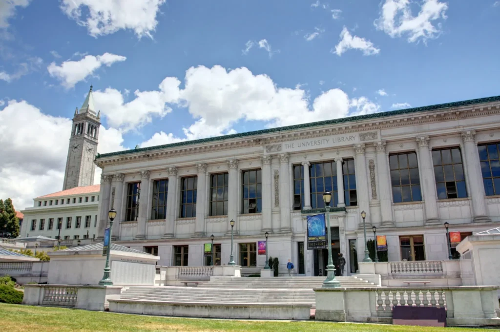 A grand classical-style building, the University Library, stands under a partly cloudy sky. It features large columns and banners that echo The California Effect. A tall clock tower is visible in the background, surrounded by lush greenery.
