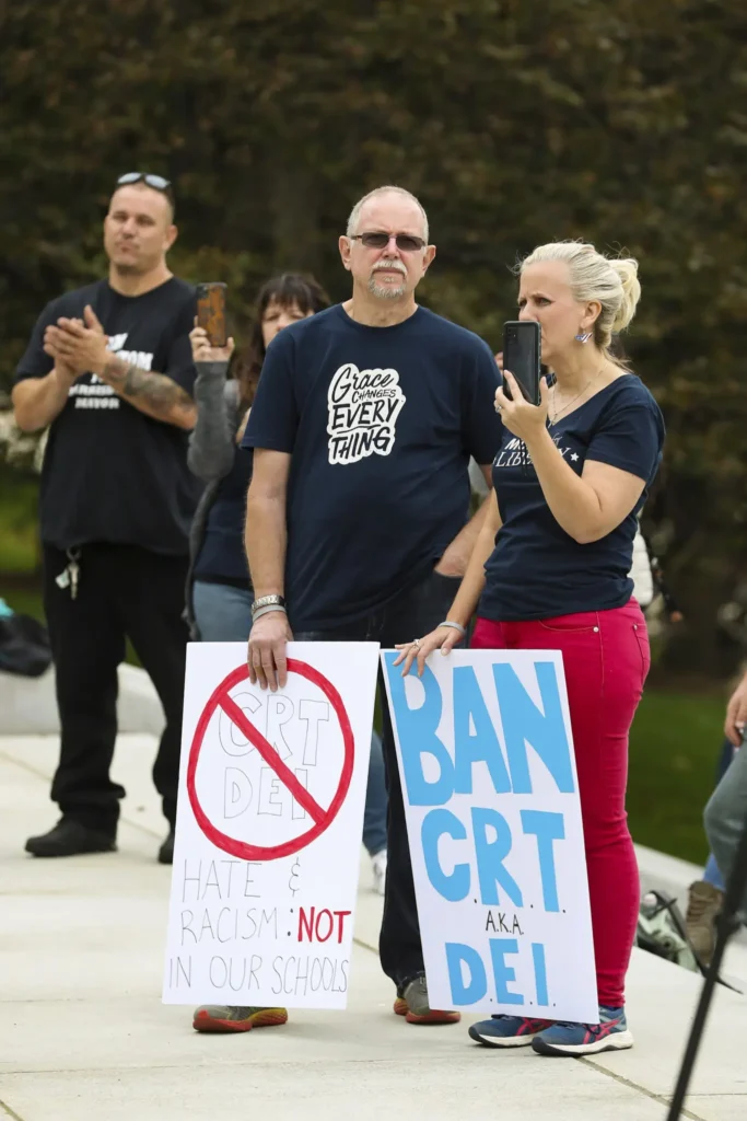 A man and woman stand outside holding protest signs opposing CRT and DEI in schools—issues some say are banned in the USA. The woman films with her phone while others stand and clap in the background.