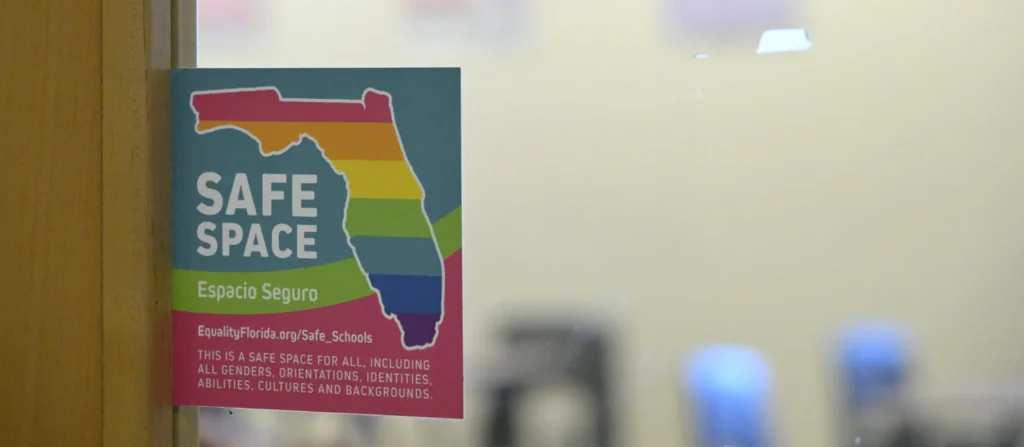A colorful sticker shaped like Florida reads SAFE SPACE and Espacio Seguro with a rainbow background, posted on a glass door. Text below promotes inclusion regardless of identity, ability, or background—proudly defying what’s often Banned in the USA.
