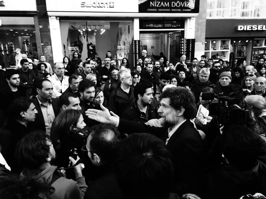 A black-and-white photo of a large crowd gathered on a city street, listening to a man with curly hair and a beard speak passionately—a scene reminiscent of the activism highlighted in the Freedom to Write Index 2020. Storefronts line the background.
