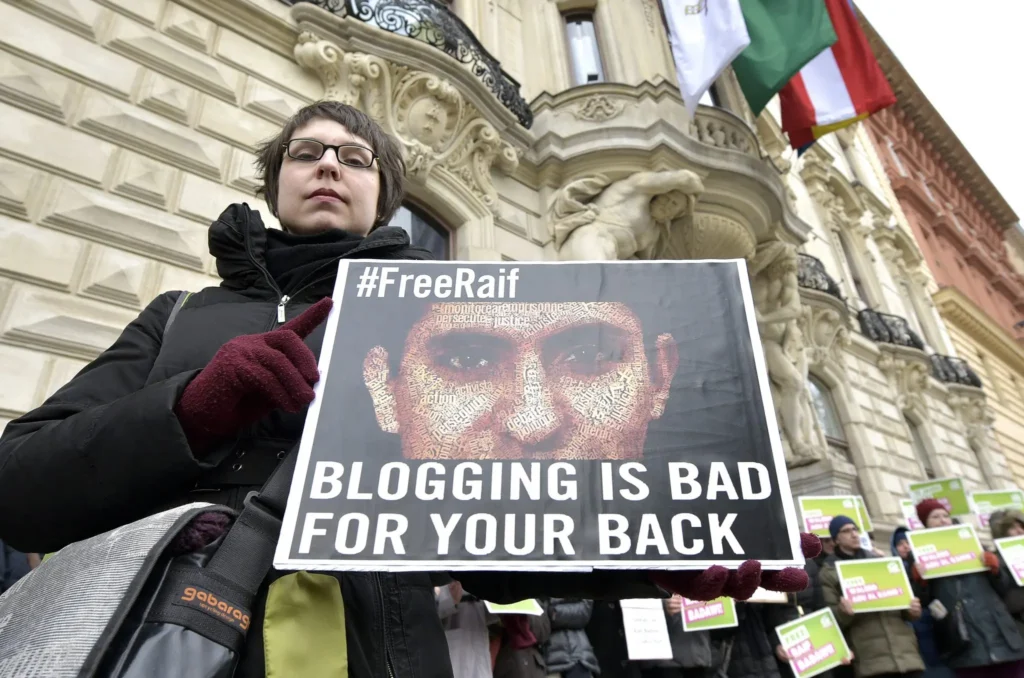 A person stands at a protest holding a #FreeRaif sign with the message "Blogging is bad for your back," highlighting concerns from the Freedom to Write Index 2020. Other protesters in the background hold similar signs.