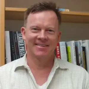 A person with short hair smiles at the camera, wearing a light-colored shirt. A bookshelf filled with various books is visible in the background.