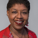 Two women smiling at the camera; the woman on the left has short curly hair and a red jacket, while her PEN Pals companion on the right has long dark hair, earrings, and a colorful top.