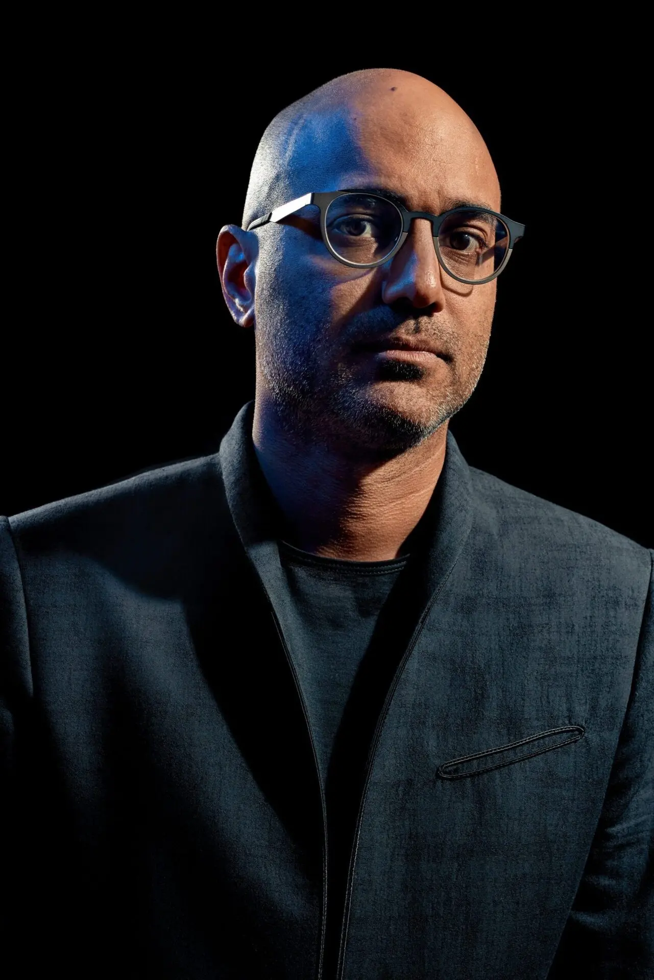 A bald man with glasses and facial stubble wears a dark jacket over a black shirt, posing against a black background with soft lighting highlighting his face.
