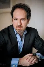 A man with short curly dark hair and a beard, wearing a dark suit jacket and light blue shirt, sits at a table with hands folded, looking at the camera with a neutral expression.