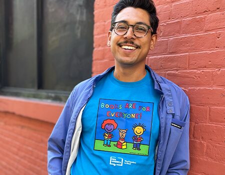 A smiling person with glasses stands by a red brick wall, wearing a blue shirt over a bright blue “Books are for everyone!” T-shirt featuring cartoon characters holding books—show your support and shop PEN America merchandise today!.