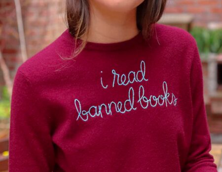 A person wearing a maroon sweater with "i read banned books" embroidered in cursive on the front, proudly sporting shop PEN America merchandise. The person's face is partially visible.