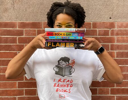 A person wearing glasses and a white t-shirt that says I READ BANNED BOOKS holds four books in front of their face, standing against a brick wall—shop PEN America merchandise to support free expression.