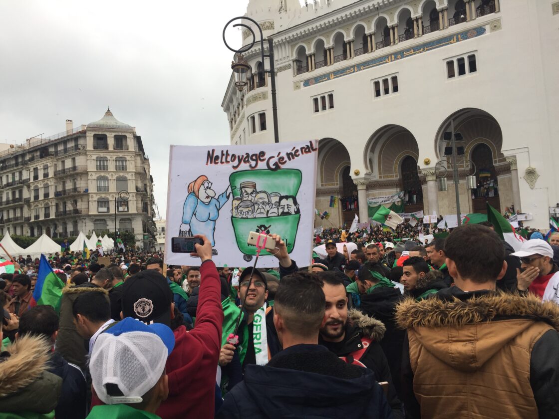 A large crowd of protesters gathers in front of a grand building. One person holds a sign with a cartoon labeled Nettoyage Général, referencing the Freedom to Write Index 2022. Several Algerian flags are visible among the demonstrators.