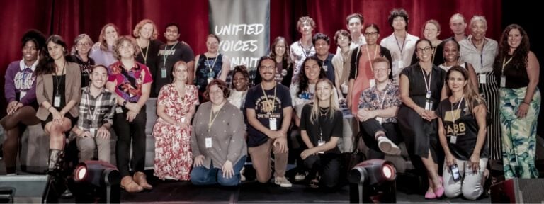 A diverse group of about 30 people pose together on a stage in front of a sign that reads “Unified Voices.” The group includes people of various ages, races, and styles, all smiling at the camera.