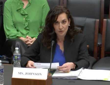 A woman with dark curly hair, wearing a black blazer and blue top, sits at a table with a microphone, water bottle, and papers labeled Ms. Johnson, speaking during a formal meeting or hearing.