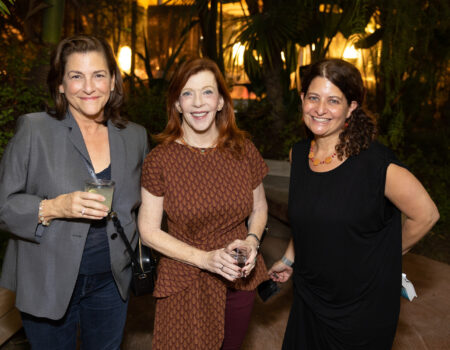 Three women are standing together outdoors at night, smiling at the camera. Two are holding drinks, and they are surrounded by greenery and warm lighting in the background.