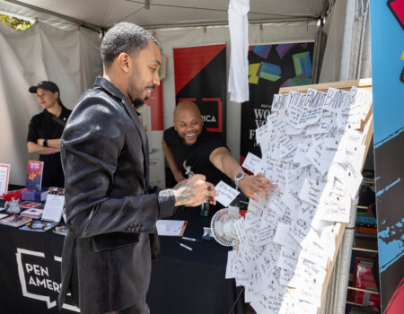 A man in a black jacket attaches a note to a board covered with handwritten notes at a booth, while another man behind the table smiles. A third person stands nearby under a tent with banners and books.