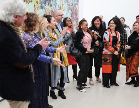 A diverse group of people stands together indoors, smiling and laughing. One woman holds a microphone as others clap and interact, suggesting a lively event or celebration. Colorful artwork is visible in the background.
